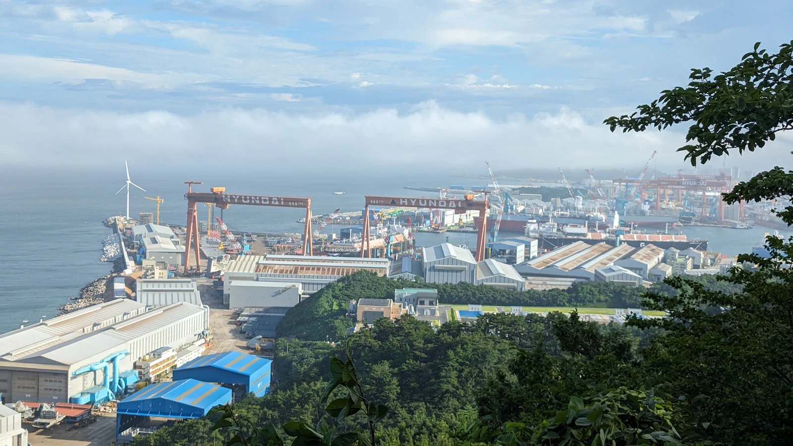 Ulsan’s Hyundai Heavy Industries shipyard viewed from Jujeon Beacon Mound on 24 July 2023. Large cranes and workshops stand beside the sea.